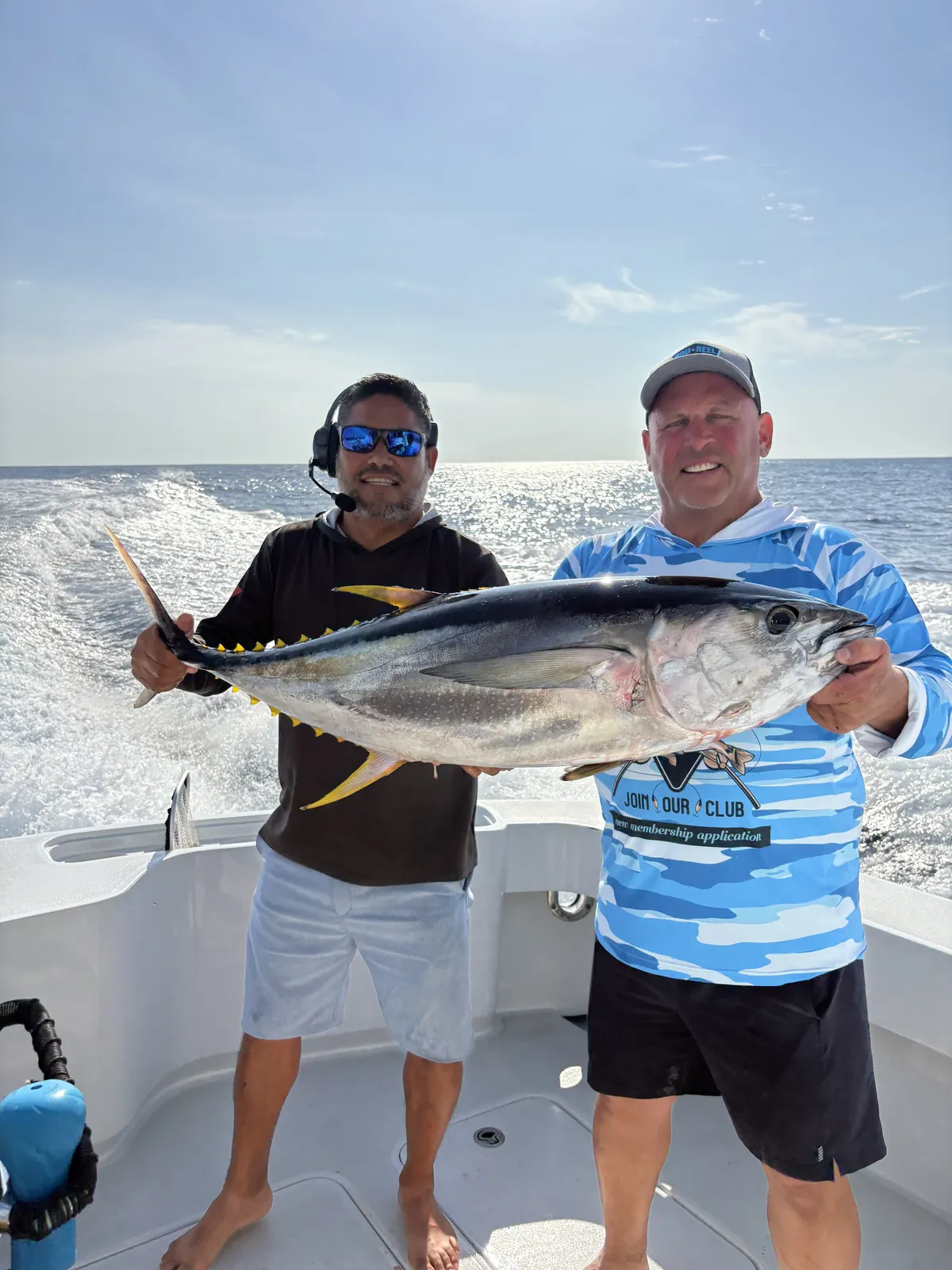 Two guests holding a yellowfin tuna on the stern with the wake behind them